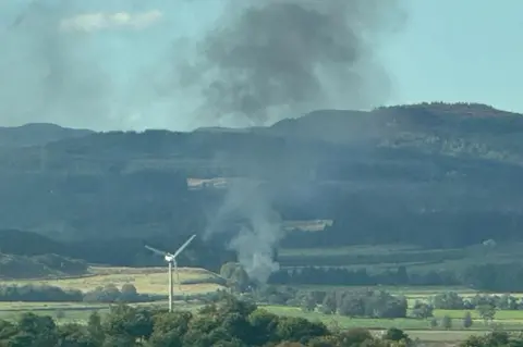 Smoke rises from the crash site. There is a wind turbine in the foreground. 