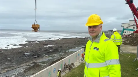 Rob Braddick is wearing a yellow hard hat and a yellow hi-vis jacket. A yellow container holding concrete is on the swing arm of the crane over the beach behind him.