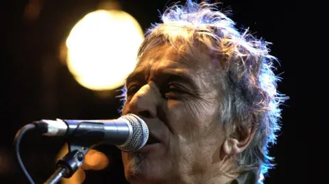 John Cale singing into a microphone. He has grey, scruffy hair and olive skin.