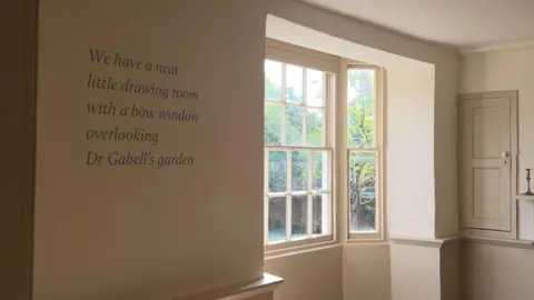 A quote in a white room with a window. the quote reads: "we have a neat little drawing room with a bow window overlooking Dr Gabells garden"