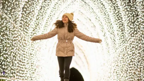 Westonbirt Arboretum A young teenage girl with long brown hair walks through an illuminated tunnel, made out of strings of fairy lights, at Westonbirt. She is smiling with her arms outstretched and wearing a quilted and belted beige coat, a beige bobble hat, dark leggings, and dark boots. 