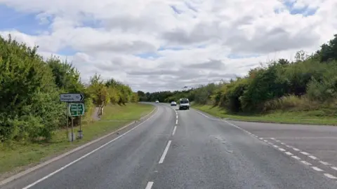 The junction of a 60mph bypass and a smaller road, both surfaced with grey asphalt and painted with standard white road markings. The bypass bends gently to the left in the distance. A van and two cars can be seen in the background. To the left, a group of three signs, one white and two green, give directions. The white sign, pointing to the right, reads "North Elkington" and gives the distance as one and a half miles. Grass verges and trees line either side of the bypass.
