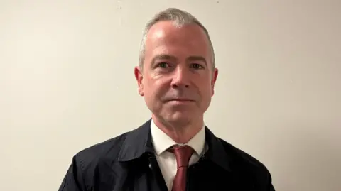 Pól Deeds has short grey hair swept back from his forehead and is wearing a dark coat, with a white shirt and red tie