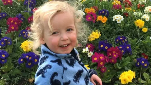 Young boy with shoulder length blonde hair, wearing a blue fleece sweater with dinosaurs, smiling standing in front of a patch of flowers.