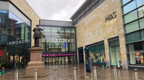 The entrance to the former M&S store in Bradford. The store front to the right of the picture is clad in light stone-coloured oblong tiles and has black M&S lettering on it. The glass-walled entrance to the shopping centre in the middle of the image carries the The Broadway