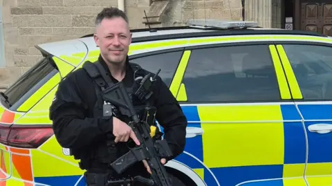 South Yorkshire Police South Yorkshire Police firearms officer PC Carl Holden wearing a black police uniform and holding a gun next to a parked police car 