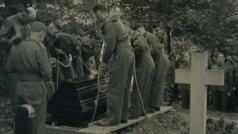 Libby MacRae A monochrome photograph from the 1940s which shows a burial ceremony in Berlin. Soldiers are lowering a wooden casket into a grave using a rope system. They stand on wooden boards beside the grave as they lower the coffin. A large stone cross is seen on the right-hand foreground.
