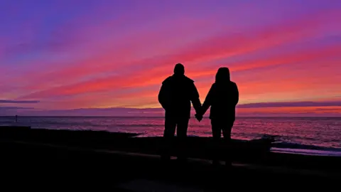 SQUIZ/WEATHERWATCHERS Two silhouetted figures hold hands while standing in front of the sea looking out at a purple coloured sunrise. 