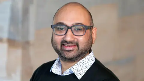 University of Essex Dr Nikhil Masters wearing a black jumper with a white shirt underneath. He has glasses with black rims on and dark stubble.