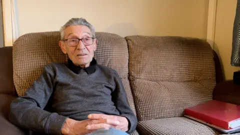 An elderly man in a grey jumper, wearing glasses and hearing aids, sits on the sofa in the lounge of his bungalow.