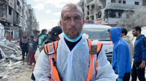 Haitham al-Homs, a man wearing orange hi-vis gear and protective forensic gear, stands in front of an ambulance in Rafah