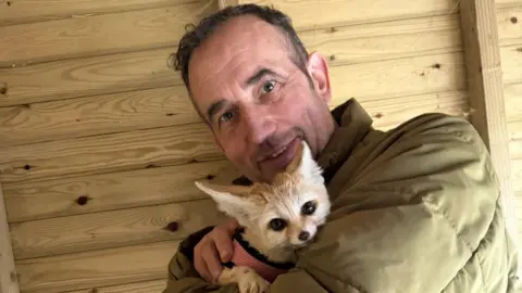Bridlington Animal Park A man with short brown hair is holding a small fennec fox and smiling. He is also wearing a green coat and is leaning against a wooden wall.