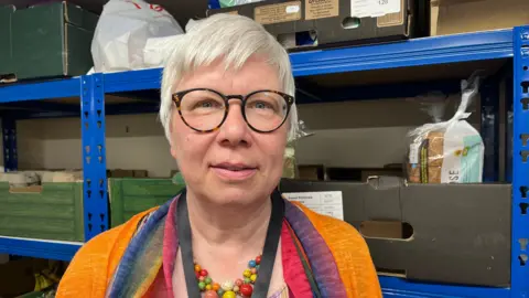 Alison looking into the camera and smiling slightly, she's wearing a bright orange cardigan with a rainbow scarf and a brightly coloured bead necklace. She's stood in front of blue metal shelving, which have cardboard boxes on with loaves of bread in.