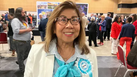 Louis Brand/BBC A woman with long brown hair wearing a creme-coloured jacket over a blue and white striped shirt with a blue scarf and a Reform UK rosette. She is smiling at the camera with people at an election count behind her.