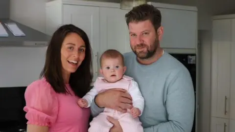 Man and woman smile widely with a baby in the man's arms.