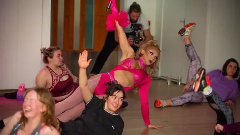 Venus Wailer Ms Wailer wearing pink tights and pink outfit leading a workout class with other women holding their legs in the air. 