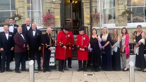Kitchener's Guest House A group of men and women standing outside Kitchener's Guest House. The men are on the left and are wearing dark suits. In the middle are three male Chelsea Pensioners, clad in bright red. On the right is a line of women in evening dress. Behind them is a yellow-brick Victorian house with bow windows on either side of its black front door.