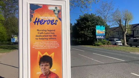 KCOM A cream phone box with an image and dedication to nurse Cath Lyon. It stands by a road with a Welcome to Hull sign on the central verge opposite.