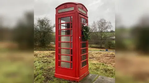 BBC A disused telephone box in the middle of a freshly cut field. There are grey slate slabs beside it, and cut grass strewn all over the ground. It is a wet overcast day and the sky is grey. The Perspex panels on the phone box are clouded and the panel at the top which reads 'Telephone' is barely legible. A Christmas wreath has been placed on the door with a white bow on it.