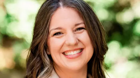 Rebecca smiling at the camera.  She has shoulder-length, wavy, dark hair and is wearing a beige blazer and a dog-tooth tweed top or dress.