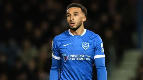 Jacob Brown stands with his arms by his side while on the pitch, wearing blue Portsmouth kit, during a match