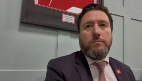 Martin Heath/BBC Pete Marland with dark hair and a beard wearing a dark purple jacket with a red Labour badge, a light brown shirt and a dark brown tie. He is sitting in front of a grey wall on which a black-framed picture is hanging.