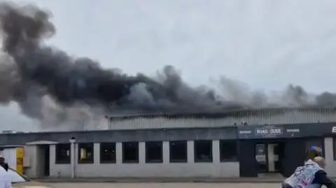 Monta Stelmahere Black smoke pours out of the roof of an industrial building. 