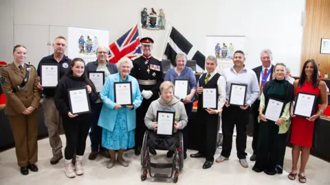 BBC A group of people holding certificates while smiling at the camera. There is a black and white Cornish flag on the right and an England flag on the left behind a man at the centre. 