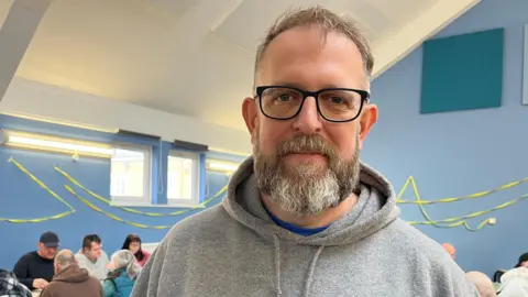 Andy Cammiss of East Durham Veterans Trust, standing in front of a blue wall dressed in a grey hoodie. He has a grey beard and hair.