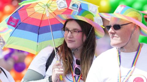 A woman with long brown hair and wearing glasses and a rainbow-patterned hat and with pink framed sunglasses tucked into her white T-shirt, holds a rainbow striped umbrella. Next to her stands a man, wearing black sunglasses, a rainbow-patterned hat, white T-shirt and a rainbow-striped lanyard around his neck.