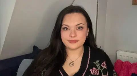 Jessica smith is a young woman. She has pale skin and long black hair that is slightly wavey. She has rosy cheeks and lips and is looking directly at the camera. She is wearing a fluffy cardigan with flowers on it, and a silver necklace with small silver hoop earrings.