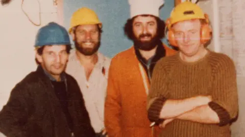 Norwegian Petroleum Museum An old photo of four men on the Kielland. Michael Fleming is on the right wearing a brown woolly jumper and yellow hard hat. To his left is a man with a thick black beard wearing orange overalls and a white chef's hat. To his left is John Murray, who is smiling with a thick and neat brown beard and yellow hard hat. The man on the far left is wearing a dark jacket and a blue hard hat.