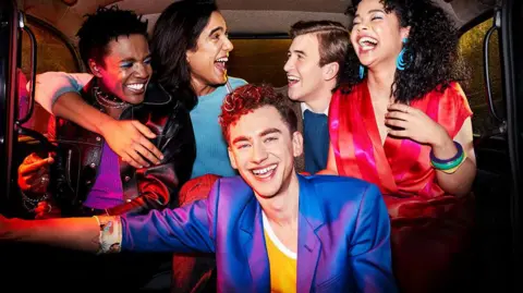 Singer and actor Olly Alexander (front) in a purple silk jacket and yellow and white t-shirt lead the cast of Channel 4's It's A Sin. He is pictured in a scene from the show in the back of a black cab with four other characters. They are all smiling.