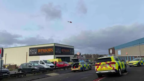 BBC Emergency services parked either side of a road. There is a car park behind and a large Wynsors store with orange and black signange. There is an air ambulance helicopter above the scene.