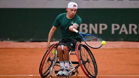 A man wearing black shorts and a green T-shirt, playing wheelchair tennis.