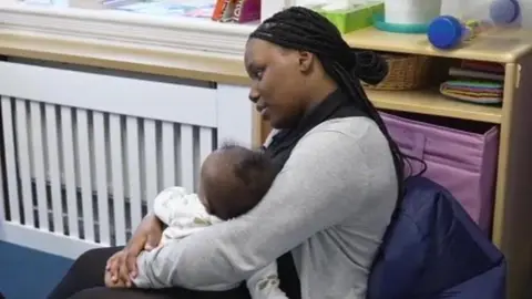 Rosabel sits on a chair whilst holding the baby 