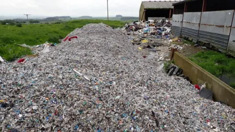 Environment Agency The image shows a large outdoor area on farmland where an extensive quantity of shredded waste has been dumped. The setting includes open countryside, farm buildings, and a wide spread of debris.
