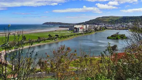 A long view of the Mooragh Lake, which sits within a green park and has an island in the middle. There are hills in the distance with blue skies with fluffy white clouds, and the beach and the sea are on the left. The Ramsey Park Hotel sits to the side of the lake.