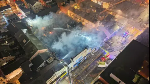 An aerial view of he building on fire with a fire engine spraying water into the area amid smoke and orange fire in one part of the building