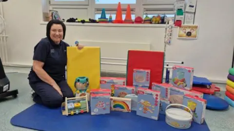 North Tees and Hartlepool NHS Foundation Trust Nicola Hewitson, who has dark hair and is wearing a navy blue polo shirt and trousers. She is kneeling next to a collection of children's toys displayed on the floor of a hospital. There are a number of boxes of toys on top of a blue mat.