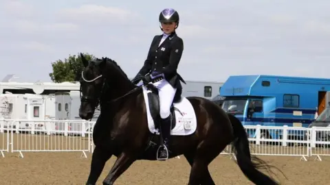 Dressage rider Alanna Clarke, riding her horse, in motion, in a paddock. The dark brown horse wears a white saddle.  The rider wears dressage uniform; white trousers,  a black riding jacket with purple trimmings,  and a purple black riding helmet.  In the background are white caravans and a blue horse box.