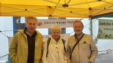 Members of Jericho Wharf Trust posing for a picture under a tent. They are 2 men and one woman. They are smiling, The trust's banner is behind them.