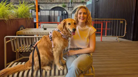 Jenn Nimmo-Smith - a woman with long blonde hair - sits on a bench in a bar's outdoor area alongside her dog Scout, a large golden retriever 