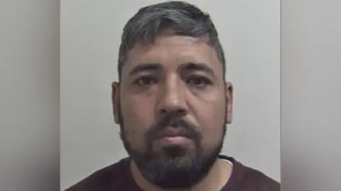 A police mugshot of a man with greying black hair and a black beard. He is wearing a brown T-shirt.