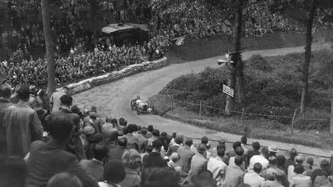 Getty Images The race captured in 1932 in a black and white image. A car is driving around an uphill bend watched by hundreds of people.