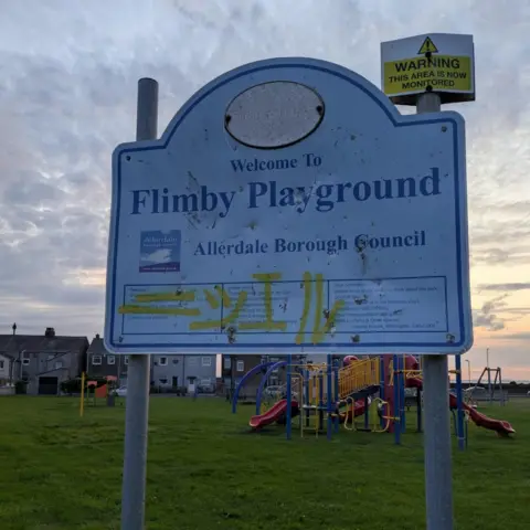 A sign, standing on the grass, at the entrance of Flimby Playground. The battered sign is covered in yellow graffiti. Behind the sign, a play area and houses can be seen.