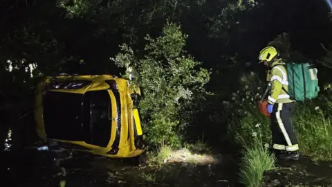 A yellow car can be seen on its side in the river, while an emergency worker in yellow high-viz is too the right with a green rucksack. 
