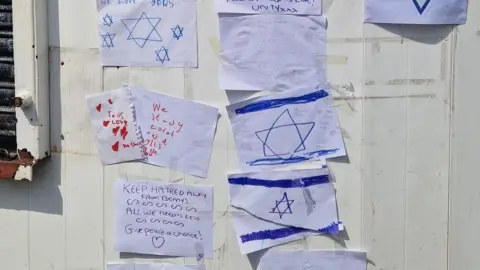 A white wall with a number of pieces of paper with handwritten messages and stars of David flags on them. 