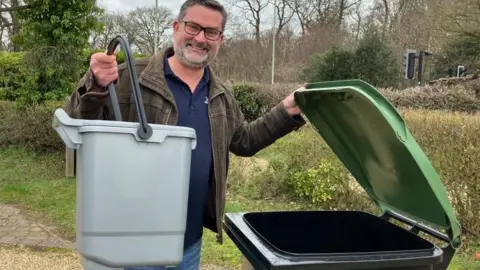 Jon Neal is standing on a gravel drive with trees and bushes in the background. He is holding a small grey caddy in one hand while standing next to a black and green wheelie bin, which he is holding open. He is wearing blue jeans, a blue jumper and a brown jacket. He is wearing glasses.