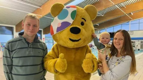 Karl Bird/BBC Parents Simon and Tammy smiling and standing either side of Pudsey, who is putting both thumbs up. Pudsey is a yellow bear wearing a colourful eye patch. Tammy is holding baby Everett who is looking at the camera and touching Pudsey's cheek. Simon is wearing a green and black striped jumper and Tammy has long brown hair and is wearing a white top with flowers on. They are standing in front of a pool.